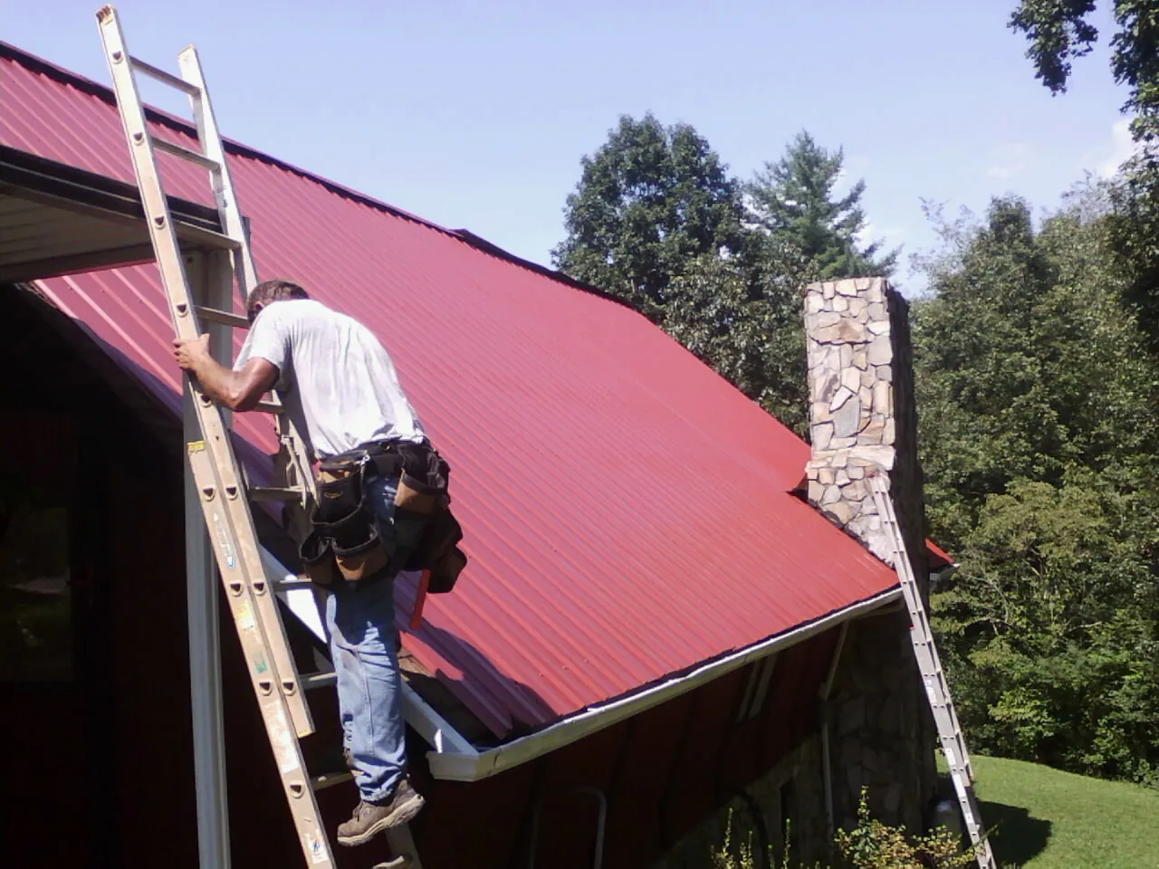 Roofer on a ladder beside a red metal roof and stone chimney — Soffit & Fascia Repair by Roofing Brothers in Iron Gate